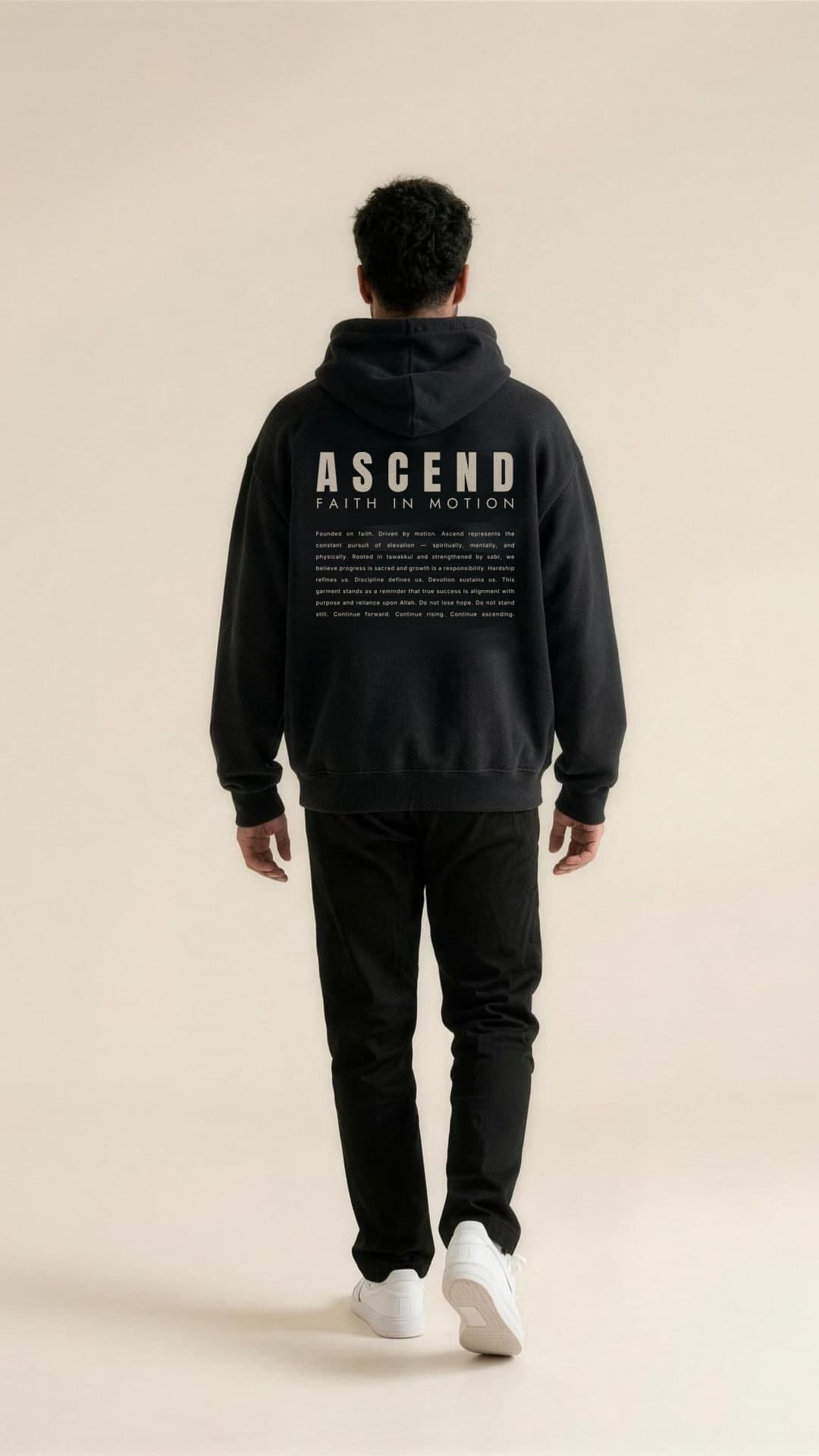 A man walks away across a cream studio in the Black hoodie, the ASCEND lockup and manifesto printed across the back